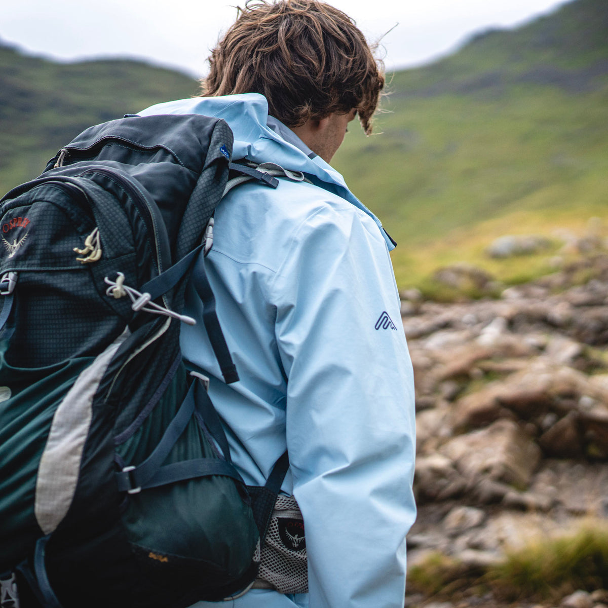 Hiker with a backpack wearing a light blue Cortazu shell jacket on a rocky mountain trail